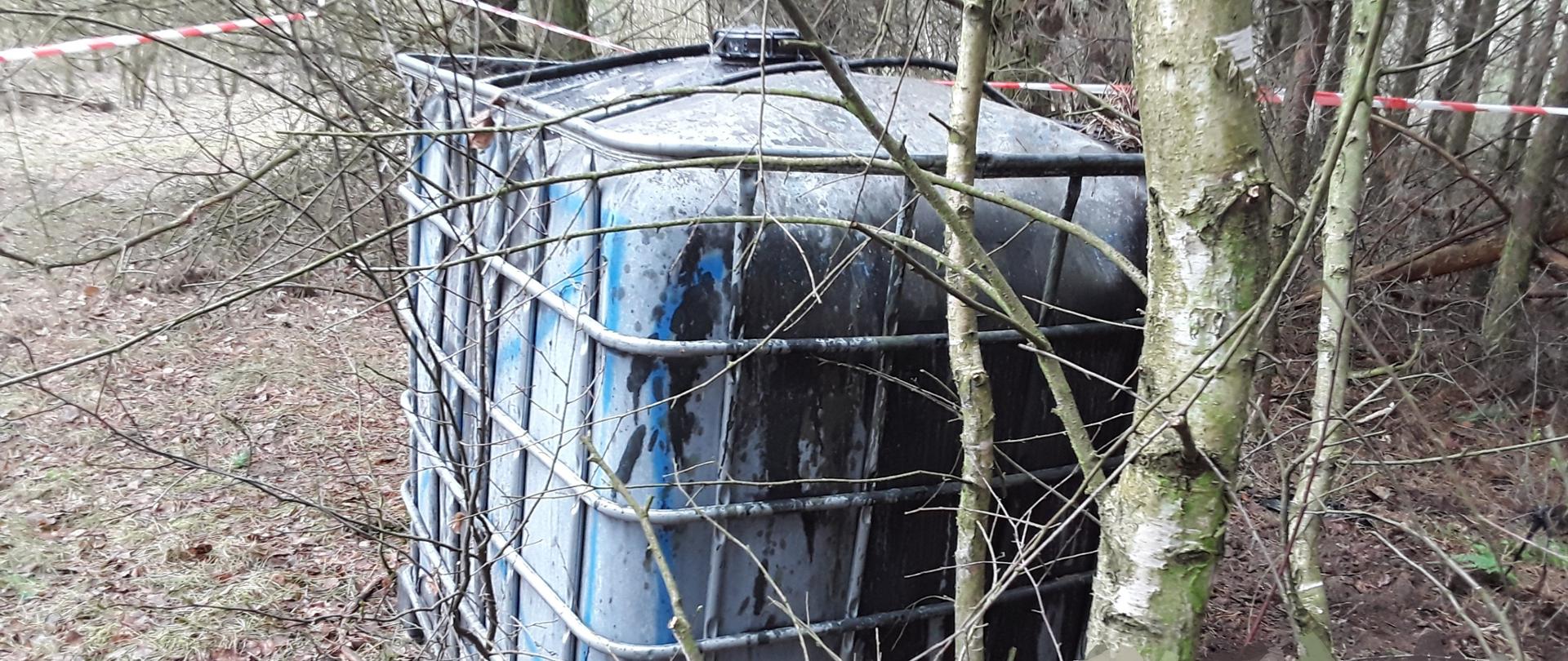 Plastikowy, biały pojemnik o pojemności 1000l stoi porzucony na nieutwardzonym terenie leśnymi.