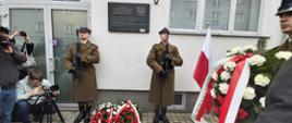 Ceremonia złożenia kwiatów pod tablicą pamiątkową umieszczoną na fasadzie budynku. 