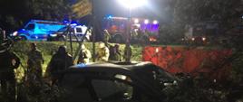 Jest pora nocna. Na zdjęciu widać uszkodzony pojazd, w dalszej perspektywie strażaków oraz wozy straży pożarnej, karetkę pogotowia oraz radiowóz.