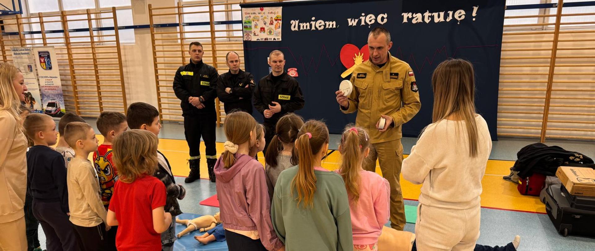 Strażacy realizujący zadania z zakresu prewencji społecznej przekazują cenne rady i wiedzę w trakcie prelekcji dzieciom w szkole na sali gimnastycznej