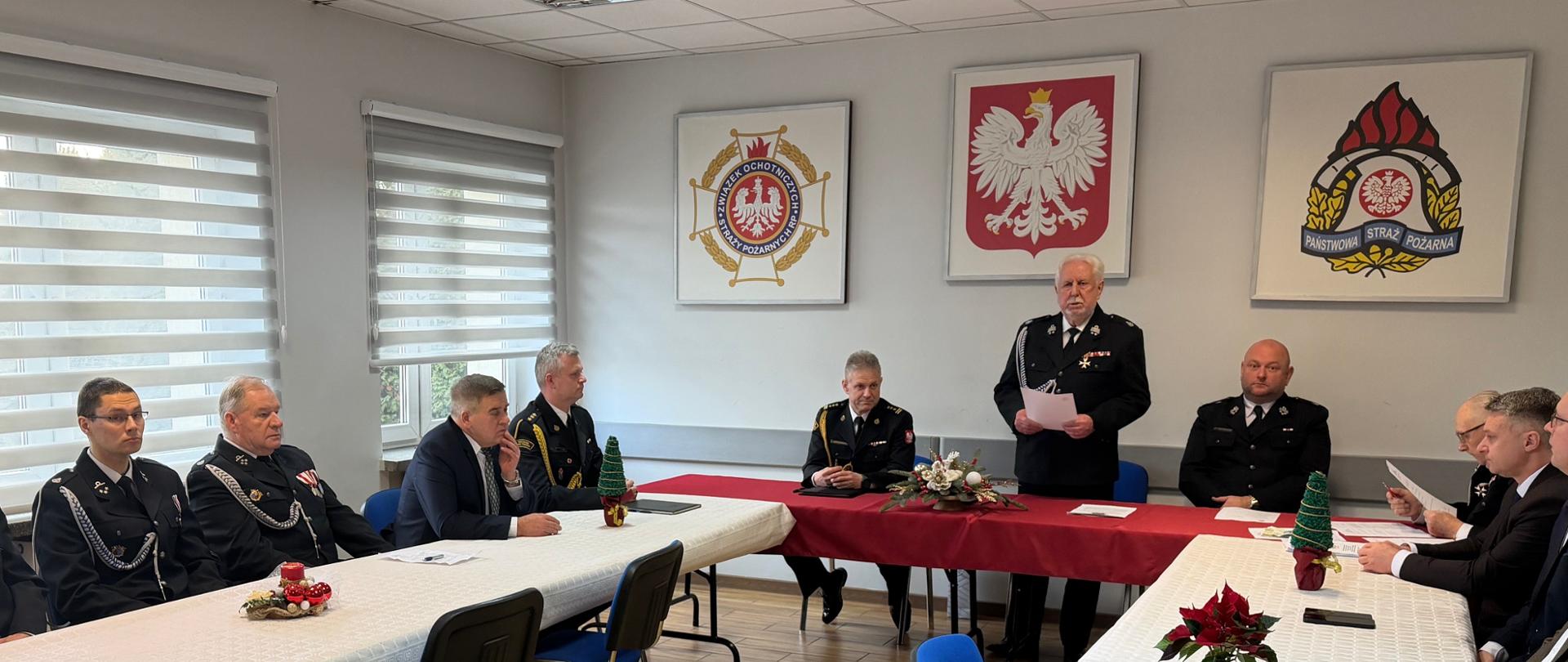 Uroczyste posiedzenie w sali konferencyjnej z udziałem strażaków w galowych mundurach oraz kilku osób w garniturach. Uczestnicy siedzą przy długich stołach nakrytych białymi obrusami, jedna osoba stoi i czyta dokument. Na ścianie wiszą godła państwowe i emblemat Państwowej Straży Pożarnej, a na stołach znajdują się świąteczne dekoracje.
