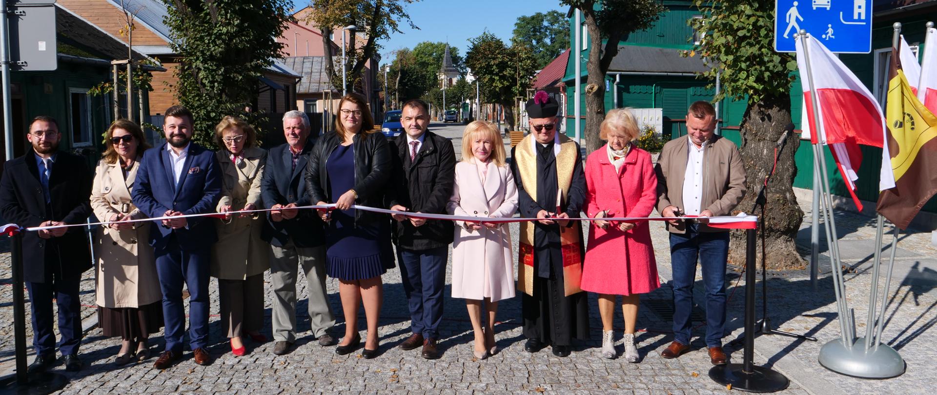 Na pierwszym planie znajduje się jedenaście osób, które stoją na nawierzchni przebudowanej drogi, przed biało-czerwoną wstęgą. W tle dwa rzędy drzew, drewniane domy, obiekty inżynierii drogowej (między innymi znaki drogowe) i piękne błękitne niebo. Uroczystości towarzyszyła piękna, słoneczna pogoda