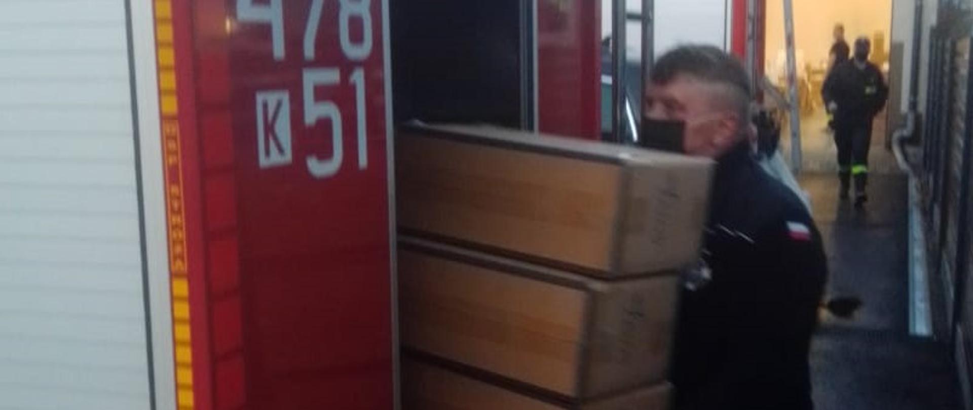 Na zdjęciu widoczny strażak ochotnik wkładający paczki ze środkami ochrony indywidualnej do samochodu strażackiego w celu dostarczenia ich do parafii.