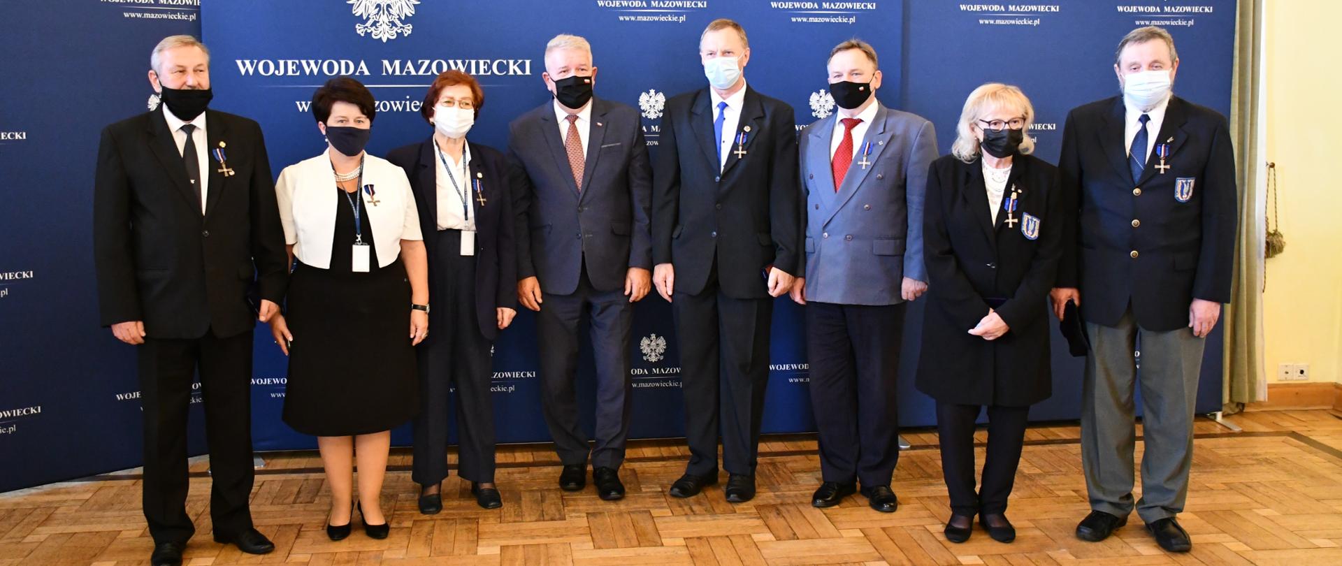 Na zdjęciu znajduje osiem elegancko ubranych osób - 3 kobiety i 5 mężczyzn. Osoby te pozują na tle granatowych ścianek wizerunkowych z Godłami Polski i napisami "Wojewoda Mazowiecki". 