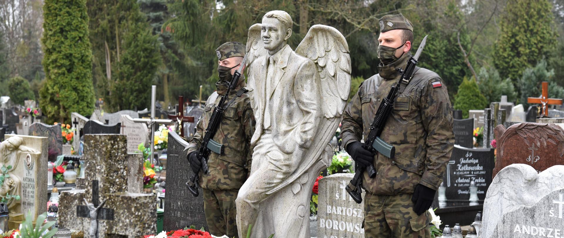 2 żołnierzy w mundurach trzyma wartę przy pomniku Bartosza Borowskiego. Na pomniku leżą biało-czerwone wiązanki kwiatów złożone przez delegacje. W tle cmentarz - inne pomniki z kwiatami i zniczami 