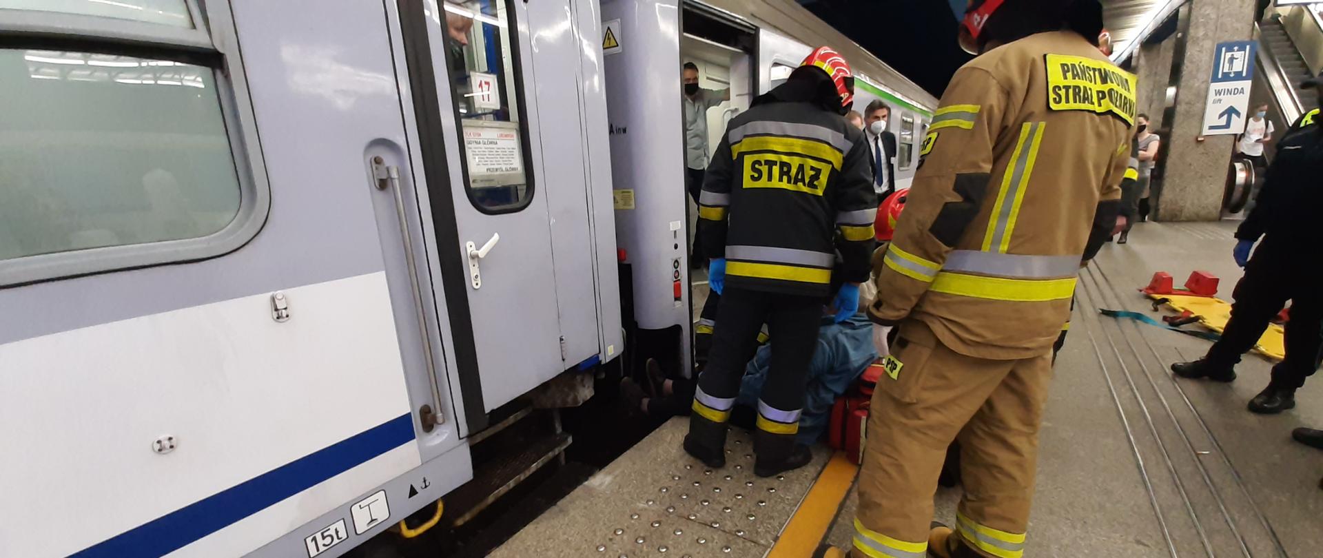 Fot. 1 Na zdjęciu na pierwszym planie widać pociąg oraz ratowników ubranych w granatowe oraz piaskowe ubrania ochronne, którzy ewakuowali kobietę z pomiędzy pociągu i peronu. Na zdjęciu znajduje się również w czarnym mundurze Policjant ,obok niego na peronie znajduję się żółte nosze typu Deska z dwoma czerwonymi zagłówkami służące do ewakuacji osoby poszkodowanej.