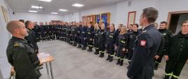 Na zdjęciu w sali konferencyjnej Komendy Powiatowej Państwowej Straży Pożarnej widoczni strażacy Państwowej Straży Pożarnej i druhowie kończący szkolenie ubrani w ubrania koszarowe w czarnym kolorze podczas uroczystego apelu kończącego szkolenie.