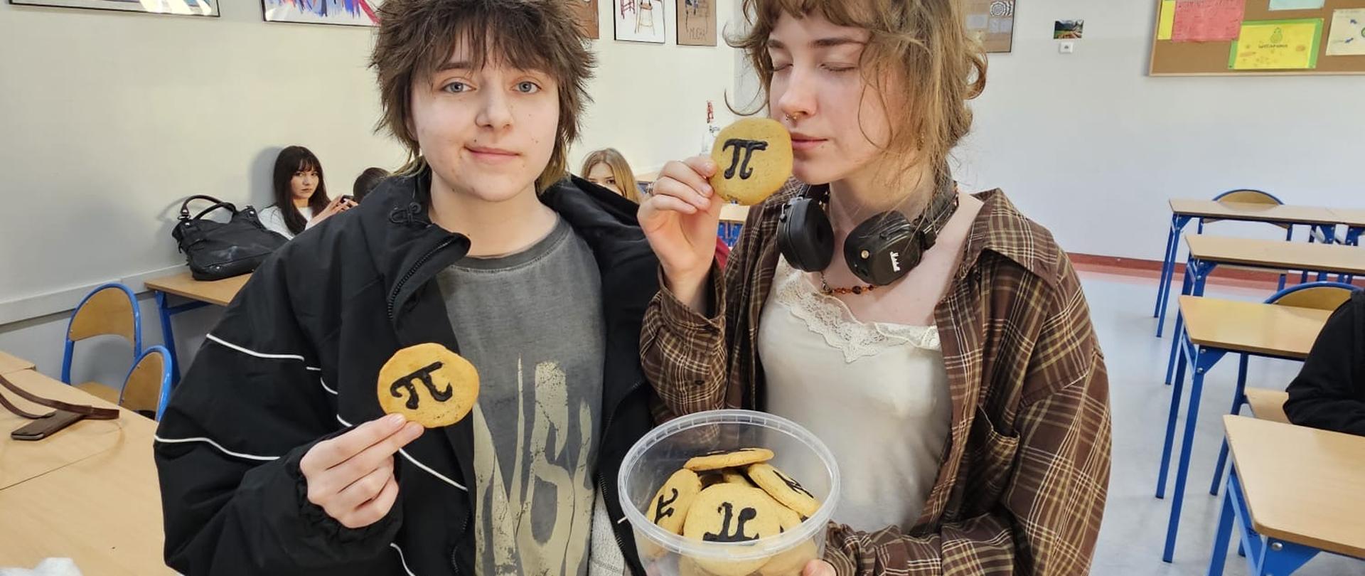 W sali od matematyki pomiędzy ławkami stoi dwoje uczniów, trzymają w ręce Pudełko z ciastkami z liczbą pi, każdy trzyma w ręce po jednym ciastku, widać że mają ochotę je zjeść