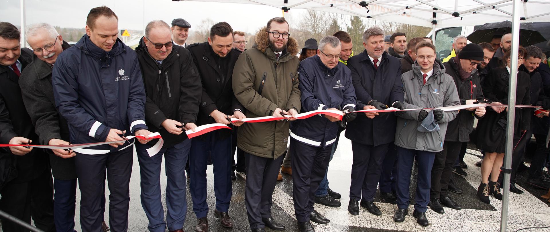 Minister Grzegorz Puda i minister Andrzej Adamczyk stoją przy biało-czerwonej wstędze, którą przecinają nożyczkami, obok nich stoją inni uczestnicy spotkania. 