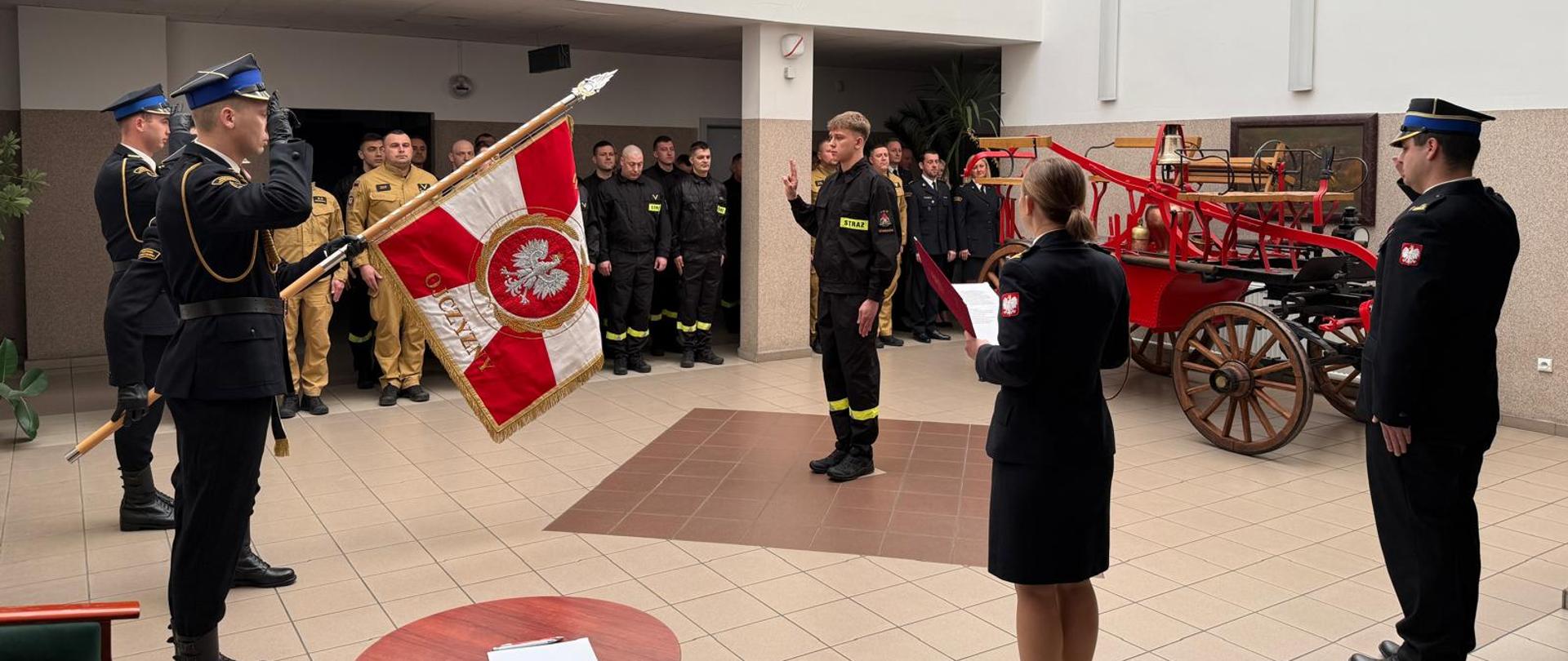 Fotografia przedstawia grupę około 20 strażaków i pracowników cywilnych Komendy, pozujących w jasnym holu budynku. Uczestnicy stoją w kilku rzędach przed zabytkowym, czerwonym elementem dawnego wozu strażackiego (sikawką konną).
W samym centrum stoi młody strażak w czarnym mundurze koszarowym, trzymający uniesioną prawą dłoń. Przed nim stoi poczt sztandarowy salutując. Po jego lewej stronie stoją strażacy w czarnych mundurach koszarowych. W środkowej części widoczni są funkcjonariusze w piaskowych mundurach służbowych. Po prawej stronie znajdują się osoby w ciemnogranatowych mundurach wyjściowych.
Wnętrze holu z jasnymi ścianami, oświetleniem wpadającym z góry przez świetlik oraz jasnymi kafelkami na podłodze.