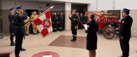 Fotografia przedstawia grupę około 20 strażaków i pracowników cywilnych Komendy, pozujących w jasnym holu budynku. Uczestnicy stoją w kilku rzędach przed zabytkowym, czerwonym elementem dawnego wozu strażackiego (sikawką konną).
W samym centrum stoi młody strażak w czarnym mundurze koszarowym, trzymający uniesioną prawą dłoń. Przed nim stoi poczt sztandarowy salutując. Po jego lewej stronie stoją strażacy w czarnych mundurach koszarowych. W środkowej części widoczni są funkcjonariusze w piaskowych mundurach służbowych. Po prawej stronie znajdują się osoby w ciemnogranatowych mundurach wyjściowych.
Wnętrze holu z jasnymi ścianami, oświetleniem wpadającym z góry przez świetlik oraz jasnymi kafelkami na podłodze.