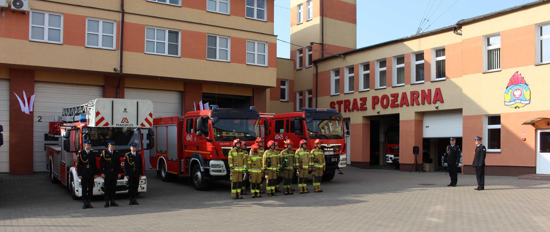 Zdjęcie przedstawia strażaków podczas uroczystego podniesienia flagi państwowej na palcu Komendy.
W tle budynek komendy.
