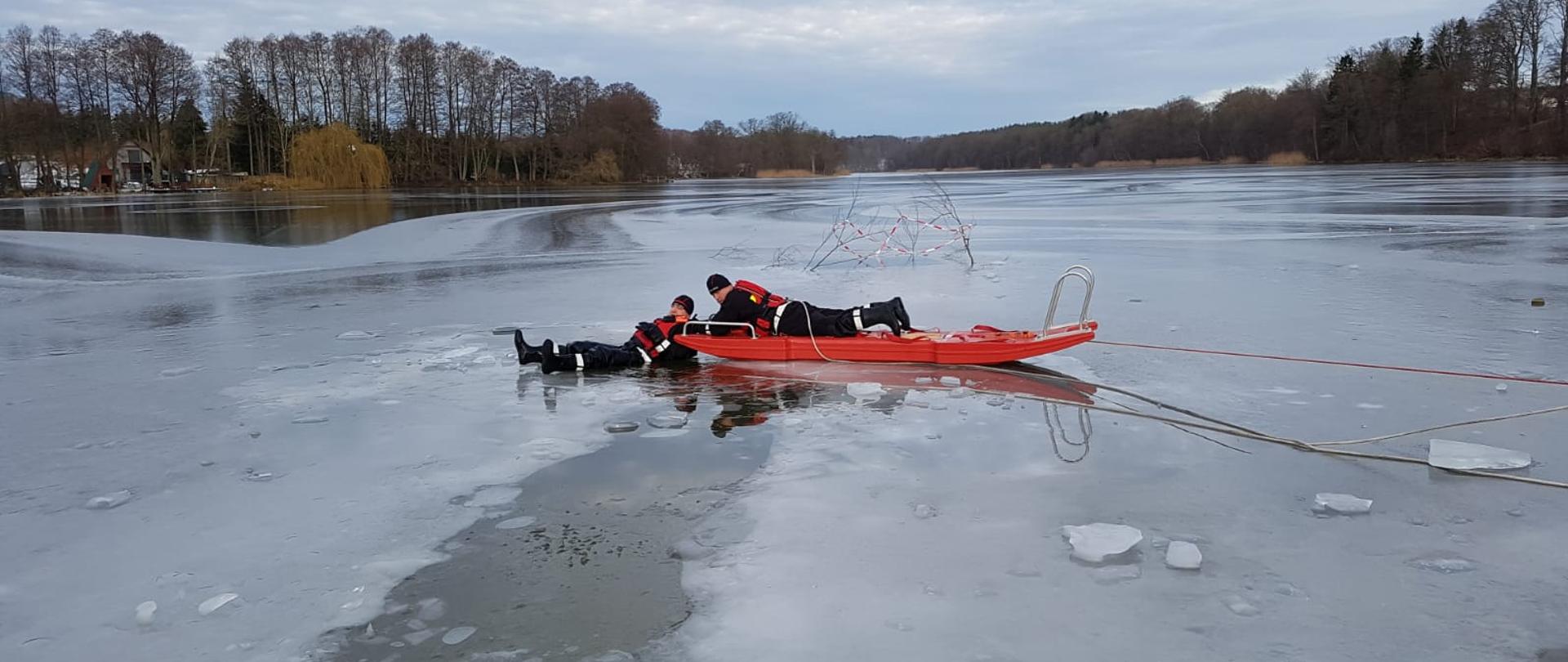 Zdjęcie przedstawia zamarznięte jezioro z widocznym podłużnym przeręblem w którym znajduje się dwóch strażaków ubranych w czarne skafandry wypornościowe. Jeden ze strażaków znajduję się w wodzie zaś drugi na czerwonych saniach wodno-lodowych z przypiętą liną. W tle widać las.