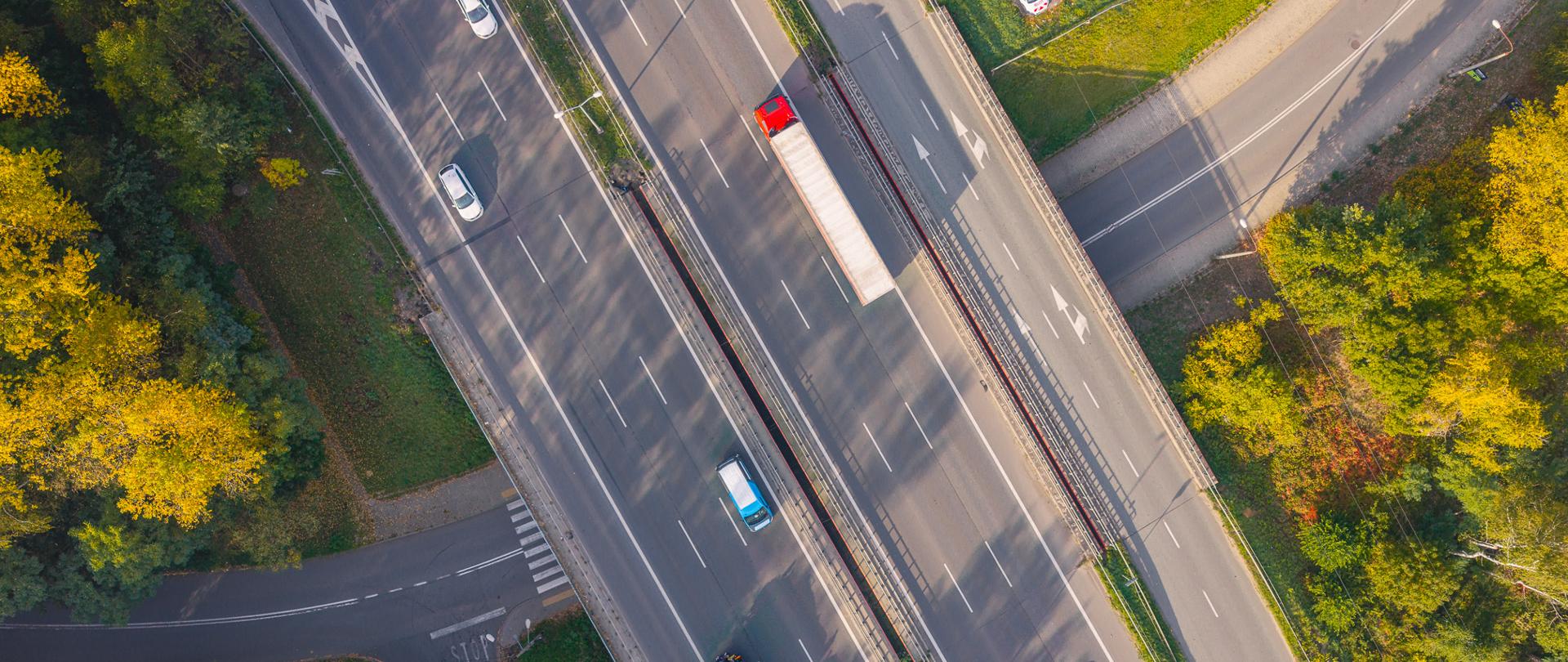 Przebiegająca przez teren otoczony drzewami w jesiennych kolorach wielopasmowa droga szybkiego ruchu łącząca Sosnowiec z Katowicami. Na jezdniach porusza się kilka pojazdów, w tym czerwony samochód ciężarowy z naczepą oraz samochody osobowe. Po prawej stronie widoczna jest jezdnia zbierająco-rozprowadzająca i zjazdy, a pod trasą - lokalna droga przecinająca teren zielony. Zdjęcie wykonano w słoneczny dzień, a długie cienie drzew padają na nawierzchnię.