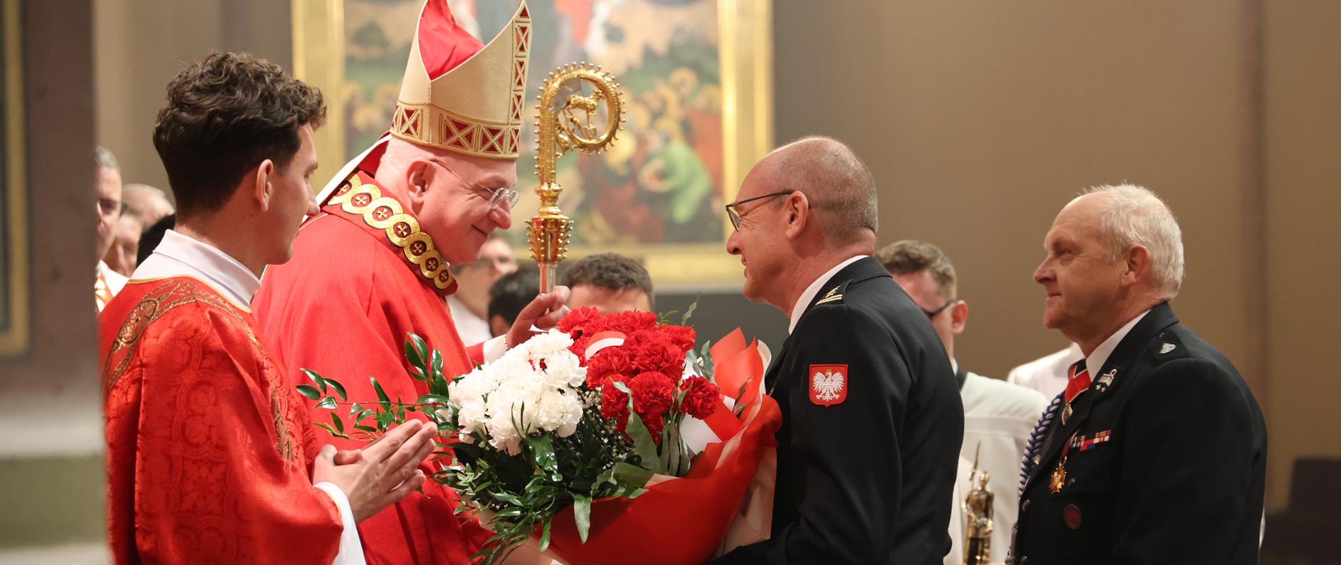 Po lewej stronie dwóch duchownych w czerwonych ornatach, po prawej dwóch strażaków w mundurach galowych. Strażak przekazuje duchownemu duży bukiet kwiatów biało-czerwonych. W tle kolorowe malowidło.