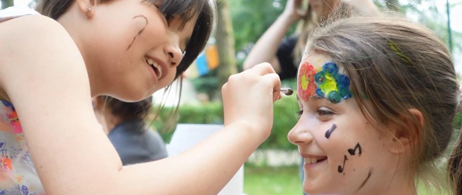 Zdjęcie kolorowe. Dwie dziewczynki. Jedna maluje na twarzy drugiej kolorowe, kwiatowe wzory. Obie dziewczynki uśmiechnięte.