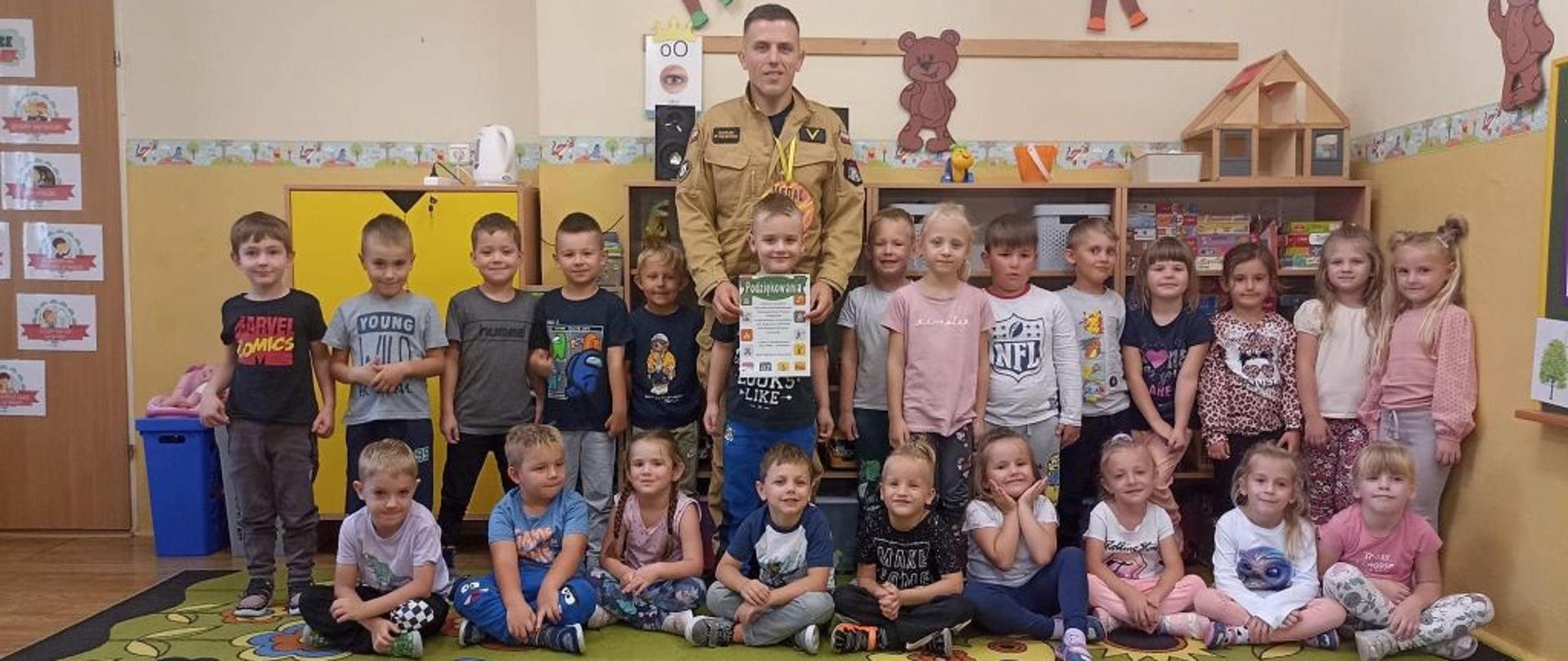 Zdjęcie grupowe dzieci i strażaka w mundurze piaskowym. Zdjęcie wykonane na sali wewnątrz budynku.