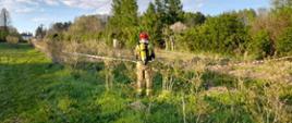 Strażak ubrany w sprzęt ochrony układu oddechowego robi pomiary stężeń niebezpiecznych gazu, który wydobywa się z uszkodzonego gazociągu. Teren jest oznakowany taśmami ostrzegawczymi. W tle widać roślinność i zabudowania.