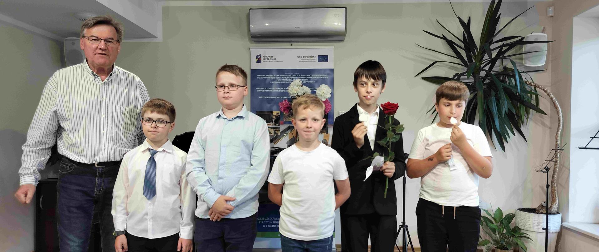 Uczniowie klasy trąbki pięciu chłopców stoją na scenie. Od lewej strony stoi z nimi ich nauczyciel. Za chłopcami baner reklamowy oraz kwiaty w doniczkach. Jeden chłopiec drugi od prawej trzyma w rekach długą czerwona różę. 