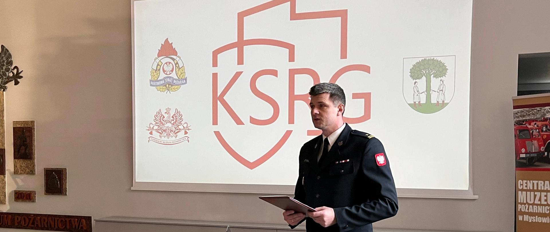 Funkcjonariusz Państwowej Straży Pożarnej wygłasza wystąpienie przy mównicy. Za nim widoczny ekran z logo Krajowego Systemu Ratowniczo-Gaśniczego oraz herbami instytucji.