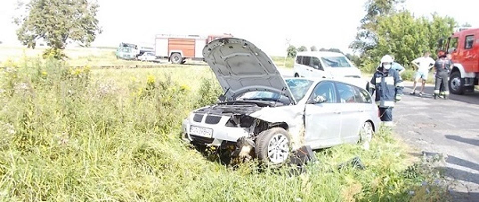 Samochód osobowy biorący udział w wypadku stoi w wysokiej trawie i zielsku. Ma otwartą maskę. W oddali na jezdni stoją samochody pożarnicze. Strażak w maseczce chirurgicznej i ubraniu bojowym stoi za samochodem.