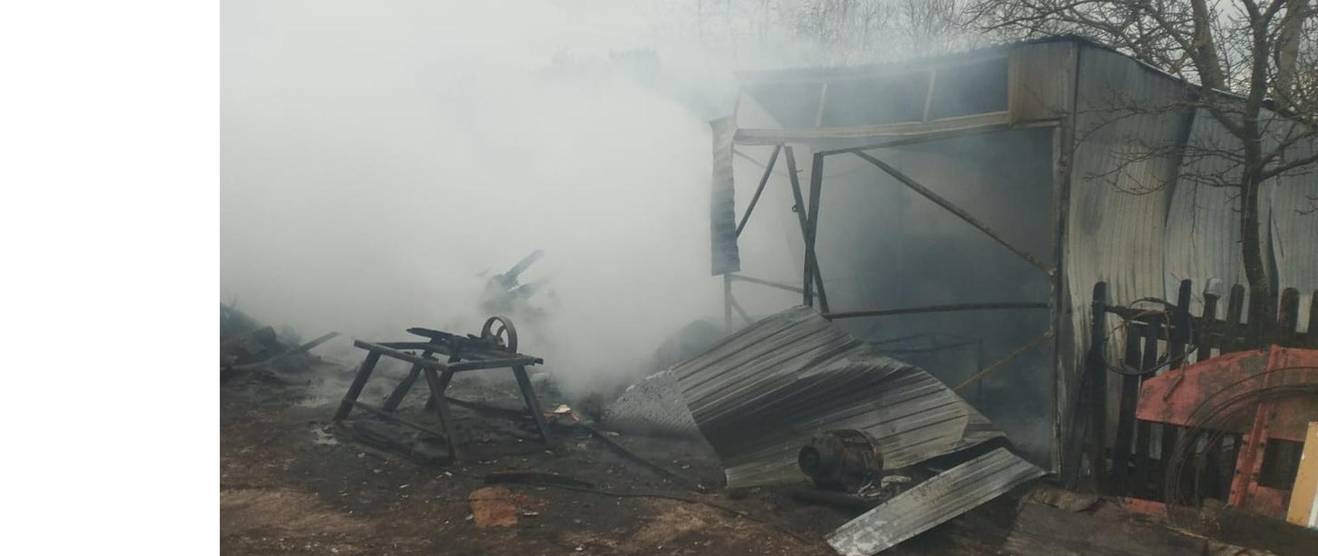 Na zdjęciu widać zniszczoną budowlę po pożarze. Konstrukcja jest częściowo zawalona, a elementy są osmolone i zniszczone. Wokół unosi się gęsty, jasny dym, który zasłania dalszą część otoczenia. Na ziemi leżą porozrzucane fragmenty spalonych przedmiotów i gruzu. W tle widać nagie gałęzie drzew i fragmenty innych budynków. 