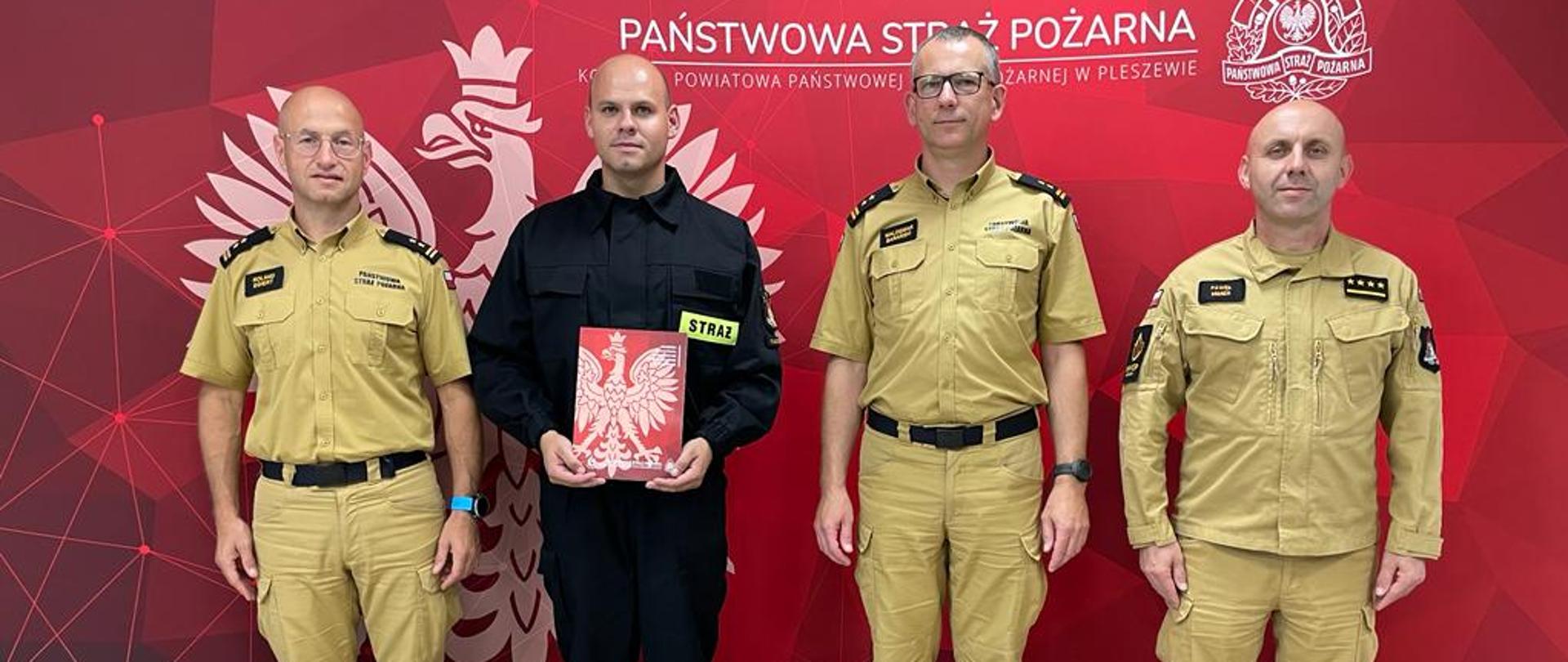 Czterech mężczyzn ubranych w mundury straży pożarnej stoi w szeregu, jeden z nich, ubrany w czarny mundur trzyma teczkę z dokumentami, za mężczyznami widać czerwony baner z białym napisem Państwowa Straż Pożarna