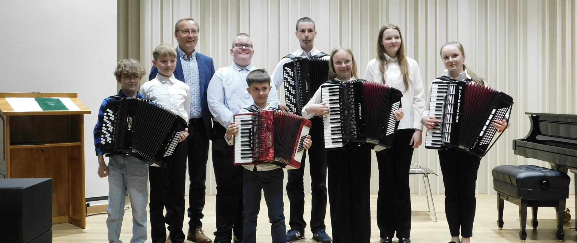 Zdjęcie wykonane podczas koncertu klasy akordeonu. Na zdjęciu grupa odświętnie ubranych uczniów, którzy trzymają akordeony. Wśród uczniów stoi mężczyzna blondyn w okularach i niebieskiej marynarce.