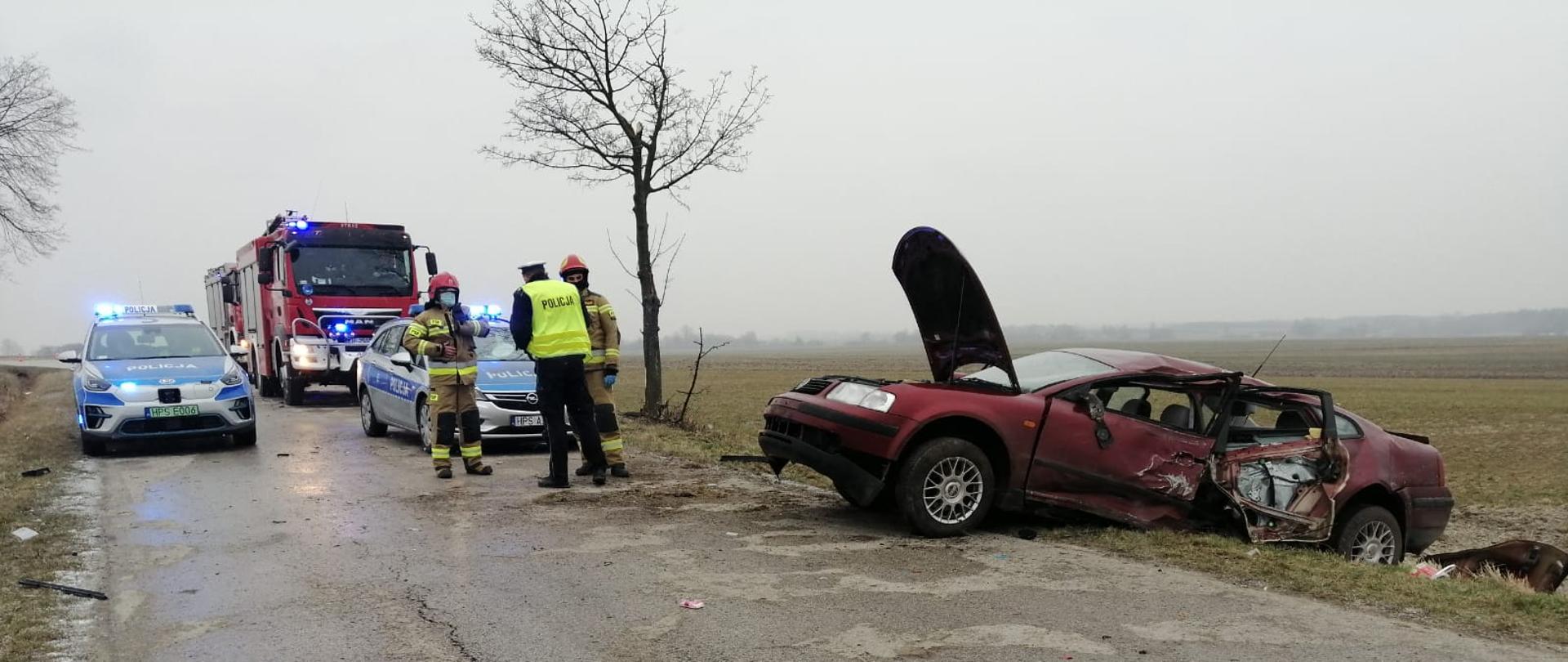 Zdjęcie przedstawia uszkodzony samochód osobowy znajdujący się częściowo na jezdni i częściowo na poboczu. Na drugim planie znajdują się dwa radiowozy policyjne oraz dwa pojazdy ratowniczo-gaśnicze z włączonymi światłami sygnalizacji alarmowej. Przed pojazdami stoją dwaj strażacy w ubraniach specjalnych oraz funkcjonariusz policji. Zdjęcie wykonane na jezdni, w tle połacie pól rolnych.