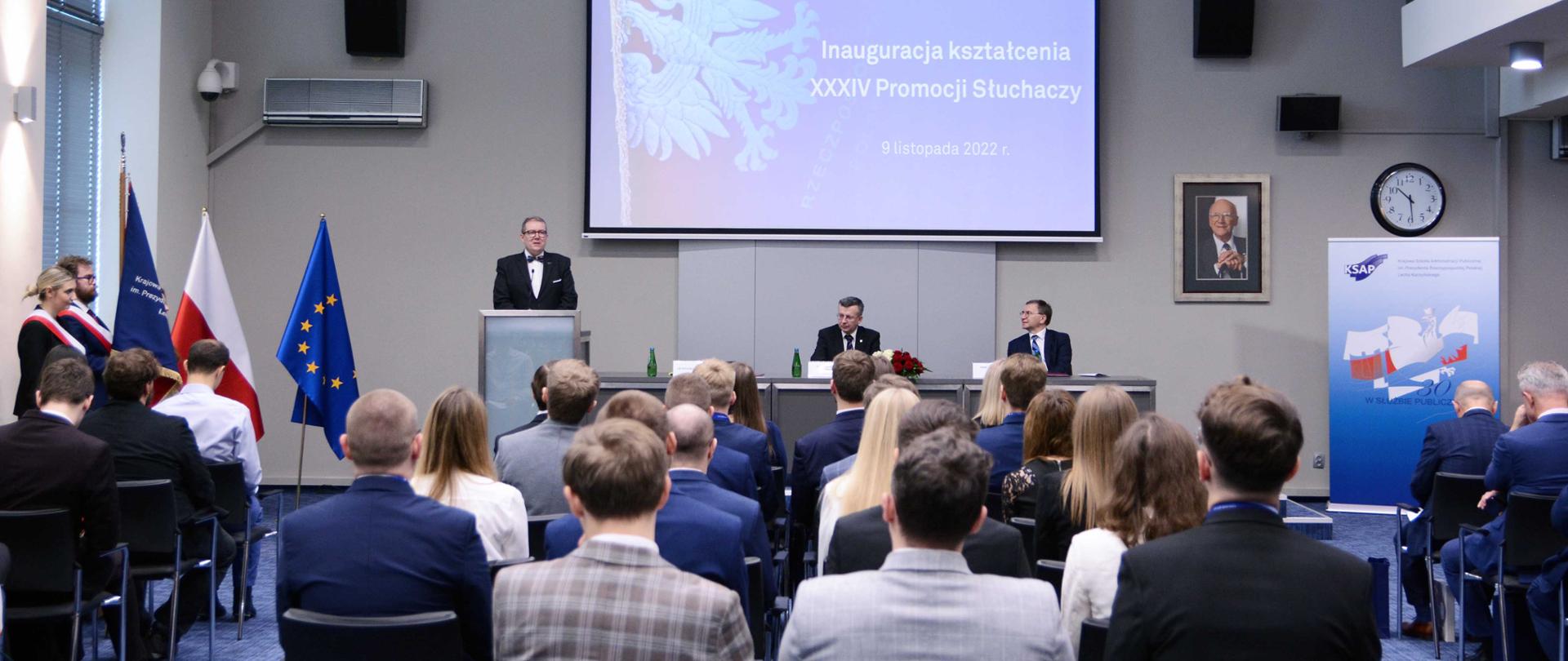 W auli siedzi kilkadziesiąt osób. Przy mównicy stoi dyrektor KSAP dr Wojciech Federczyk. Przy stole prezydialnym siedzi szef służby cywilnej Dobrosław Dowiat-Urbański i jeden z wykładowców KSAP. Po lewej stronie poczet sztandarowy KSAP oraz flagi Polski i Unii Europejskiej. 
