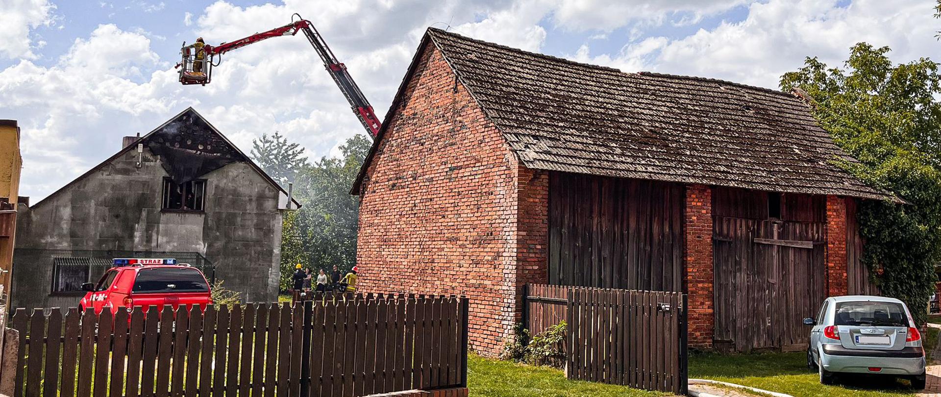 Samochód operacyjny stoi przed budynkiem, w którym miał miejsce pożar. Obok rozwinięta linia gaśnicza po trawie. Po prawej stronie ceglana stodoła, za nią widać podnośnik strażacki w trakcie działań. W tle zabudowania miejskie.