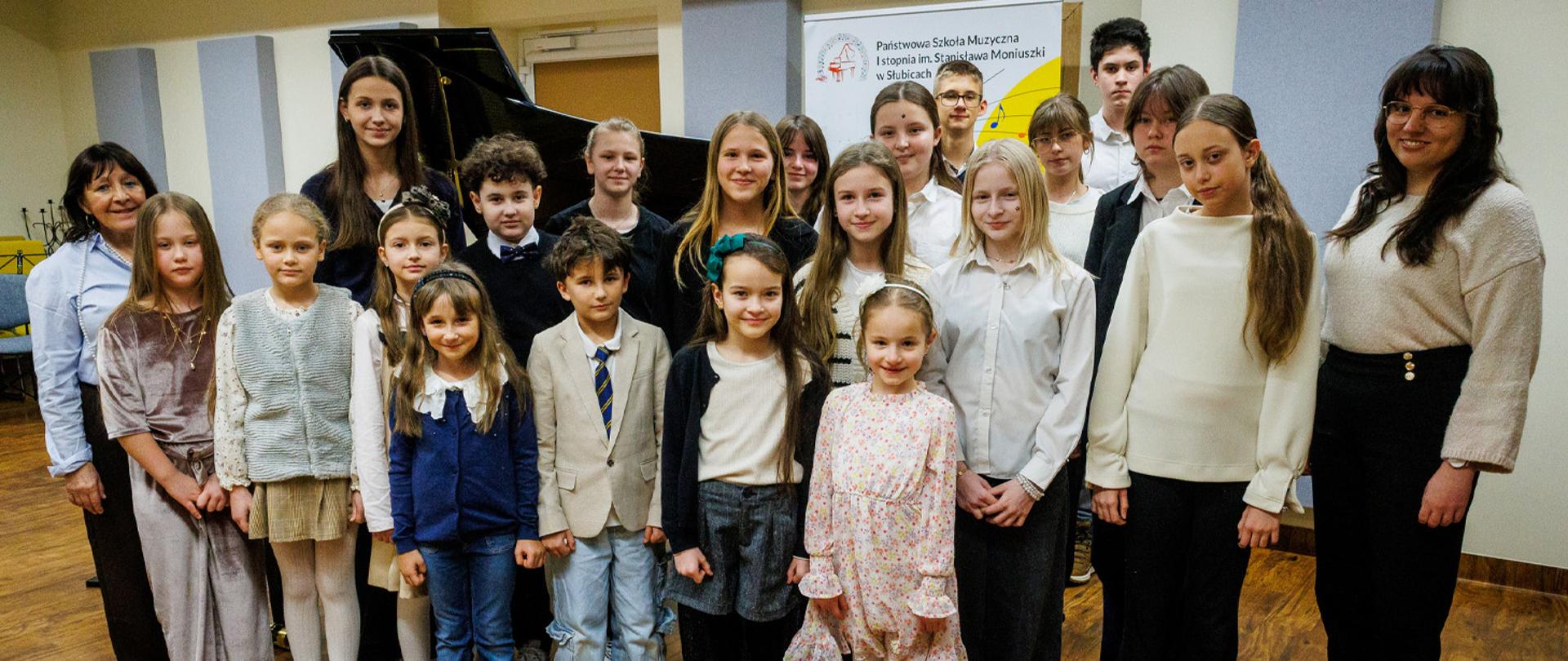 Dwie nauczycielki i kilkunastu uczniów stoją w auli szkolnej, za nimi stoi fortepian i baner szkolny.