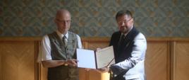 Wiceminister Rzymkowski stoi obok mężczyzny w kamizelce, obaj trzymają otwartą teczkę z dokumentem.