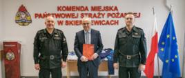 
Na zdjęciu dwóch strażaków w mundurach koloru czarnego oraz mężczyzna w garniturze koloru granatowego na tle czerwonego napisu Komenda Miejska Państwowej Straży Pożarnej w Skierniewicach, loga psp, flag polski i unii europejskiej.