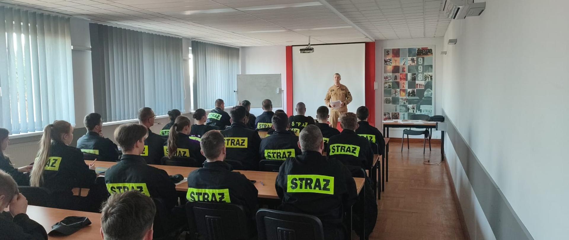 Strażący w mundurach siedzą na sali szkoleniowej. Strażak prowadzi zajęcia.