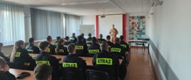 Strażący w mundurach siedzą na sali szkoleniowej. Strażak prowadzi zajęcia.