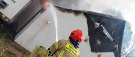 Na zdjęciu widać strażaka lejącego wodę a dach palącego się budynku. Zdjęcie wykonane w dzień. 