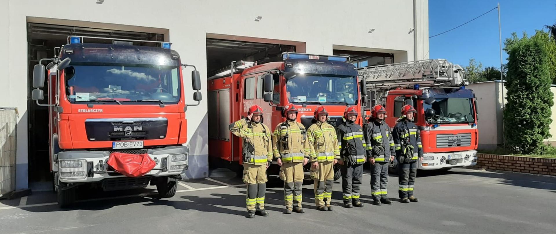 strażacy stoją przed jednostką ratowniczo - gaśniczą dowódca oddaje honor, w tle pojazdy ratowniczo - gaśnicze 