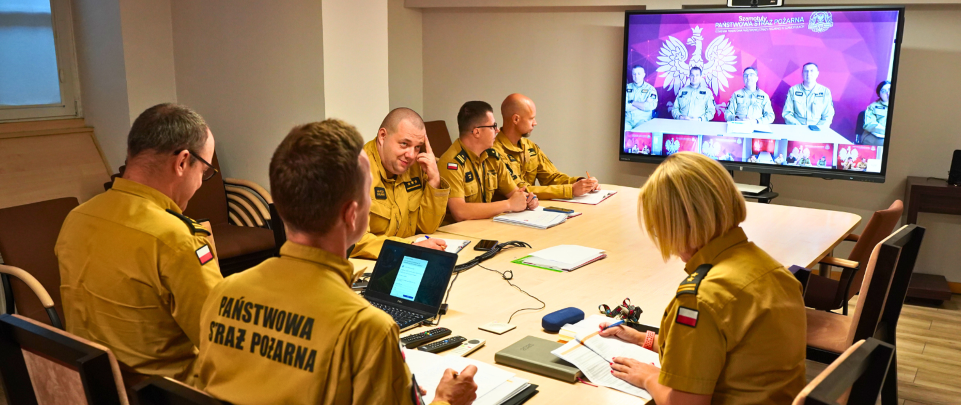 Strażacy siedzą za stołami w sali konferencyjnej i wysłuchują mówców podczas połączenia wideokonferencyjnego.