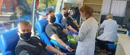 Zdjęcie przedstawia trzech strażaków w czarnym koszarze dowódczo-sztabowym siedzących w busie na fotelach do oddawania krwi. Strażacy mają podpiętą aparaturę do pobierania krwi. Wewnątrz busa są dwie pielęgniarki w białym kitlu. 