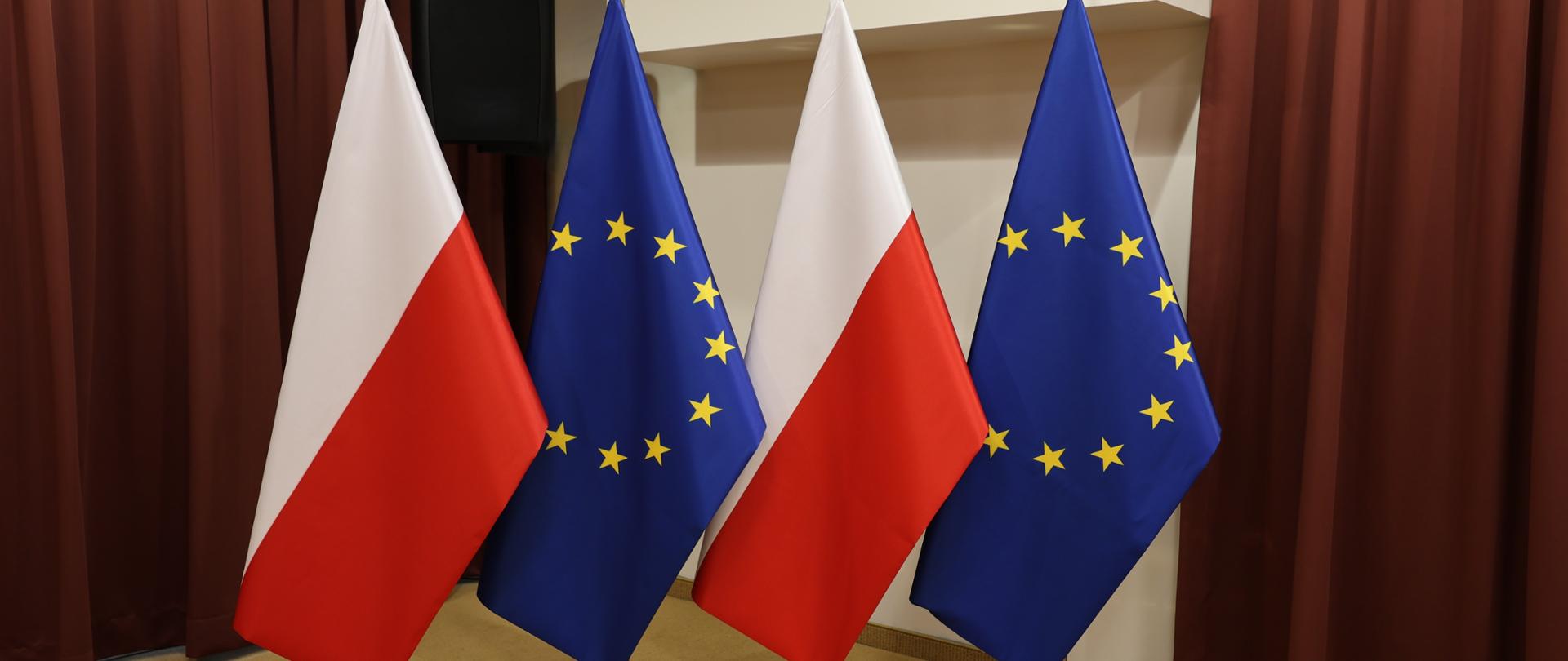 Flagi Polski i Unii Europejskiej na tle kremowej ściany oraz bordowych kotar. 