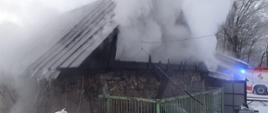 Zdjęcie przestawia budynek murowany kryty blachą z którego wydobywa się dym w tle jest widoczny pojazd pożarniczy