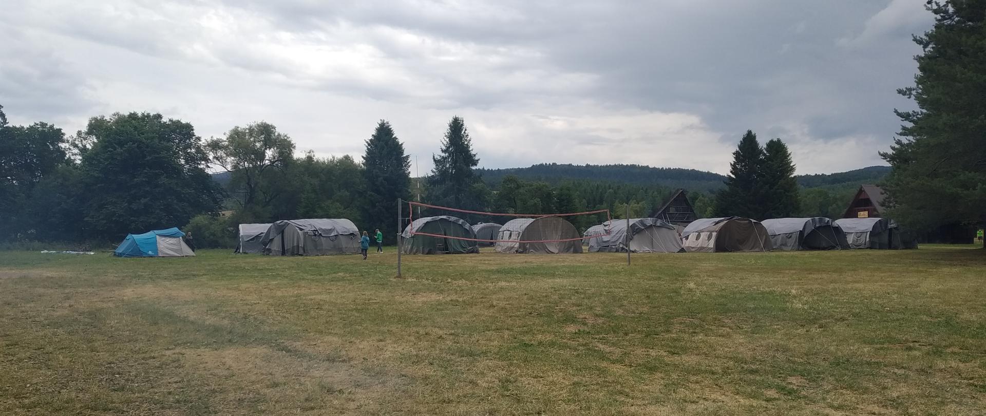 Zdjęcie przedstawia namioty zlokalizowane na łące. W tle domki wypoczynkowe oraz las. Na pierwszym planie boisko do siatkówki.
