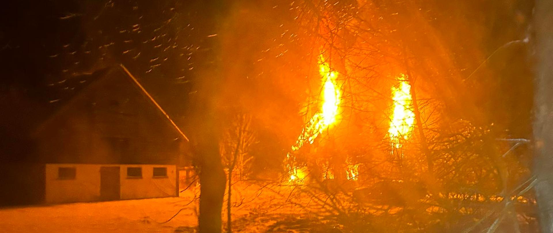 Pali się budynek mieszkalny, obok niego stoi budynek gospodarczy. Pora nocna