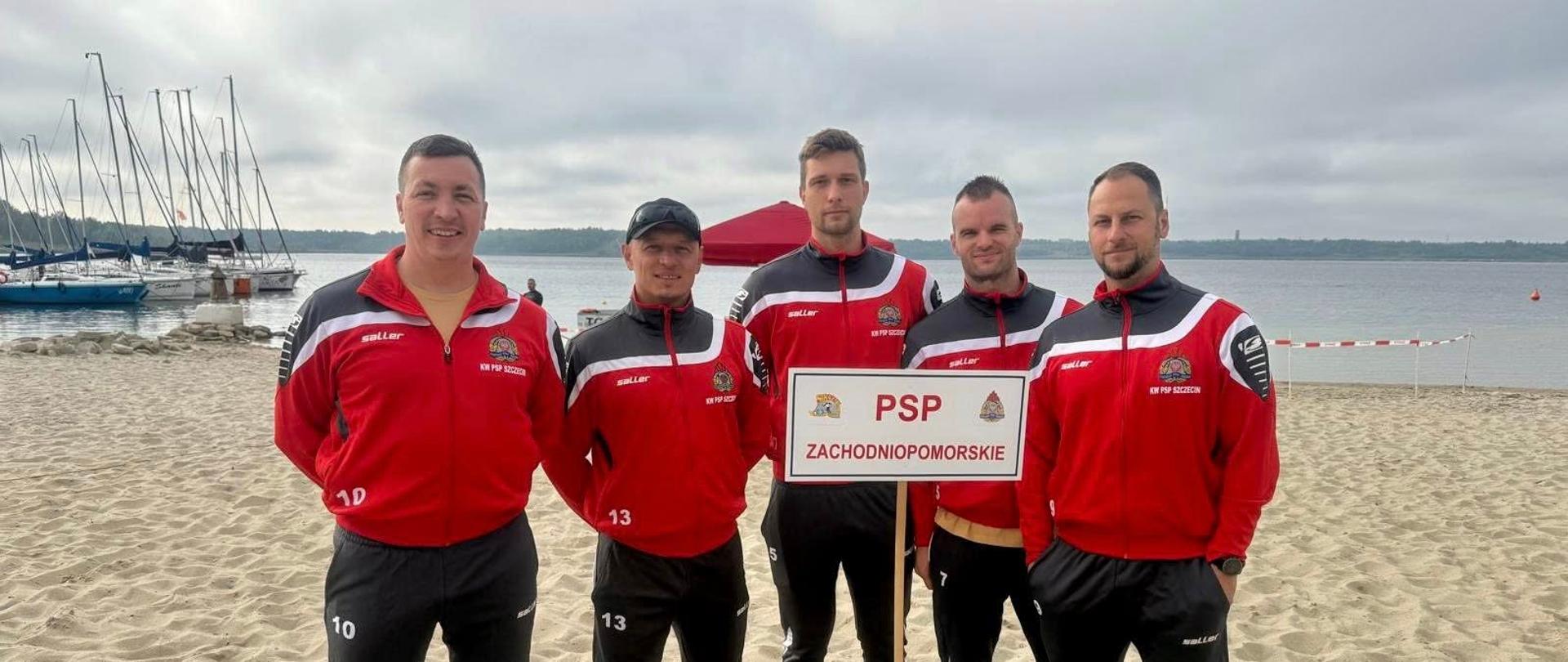 Na zdjęciu widać pięciu mężczyzn stojących na piaszczystej plaży nad jeziorem lub zalewem. W tle znajdują się jachty zacumowane przy pomoście oraz woda z linią lasu na horyzoncie. Wszyscy mężczyźni mają na sobie jednakowe stroje sportowe: czarne spodnie dresowe i czerwone bluzy z herbem Państwowej Straży Pożarnej oraz napisem „KW PSP Szczecin”. Środkowy mężczyzna trzyma tablicę z napisem: „PSP ZACHODNIOPOMORSKIE”, wraz z herbem województwa. Czterech z nich ma na nogach sportowe buty, a jeden stoi boso. Wszyscy patrzą w stronę aparatu i pozują do zdjęcia.
