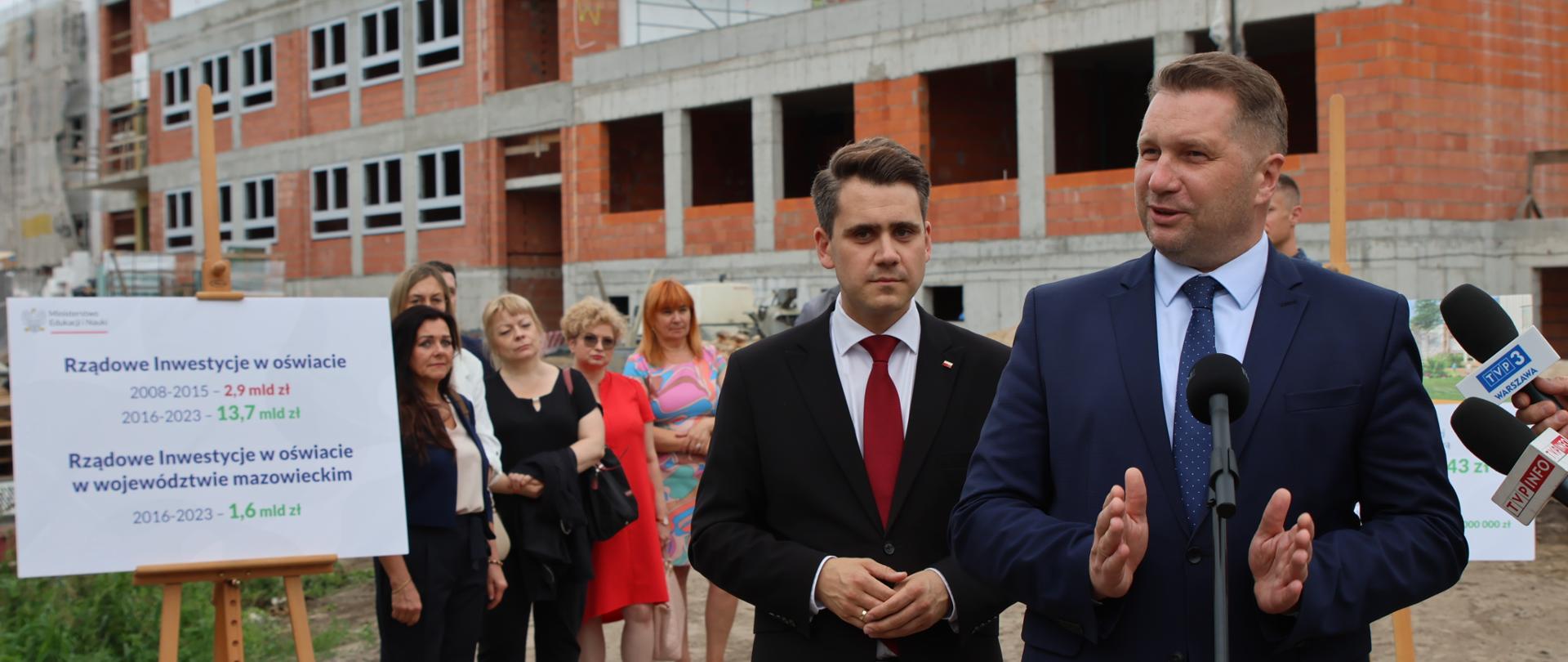 Minister Czarnek stoi przy mikrofonie, za nim mężczyzna w garniturze. W tle widać budynek w trakcie budowy i grupę kobiet.