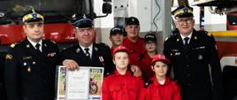 Na zdjęciu widać 5 dzieci z MDP wraz ze swoim opiekunem druhem z OSP z powiatu opatowskiego, którzy pozują do zdjęć z nadbryg. Krzysztofem Hejdukiem oraz nadbryg. Krzysztofem Cioskiem. Dru z OSP trzyma w ręku decyzję o wręczeniu promesy dla MDP na kwotę 6000 zł. Zdjęcie zrobione na tle samochodów pożarniczych w garażu JRG 1 KP PSP w Ostrowcu Św.