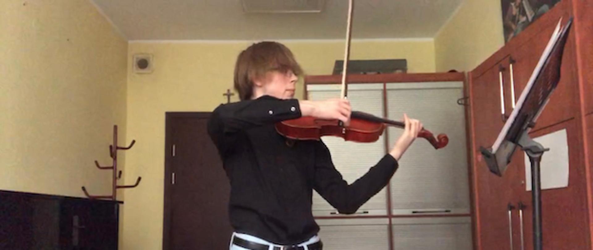 Uczeń w klasie altówki, trzyma altówkę i smyczek, ubrany w czarną koszulę i jasne spodnie, obok pianino, szafa.