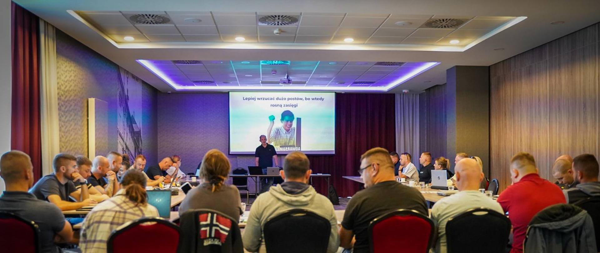 Uczestnicy konferencji siedzą przy stole, słuchając wykładu i oglądając prezentację