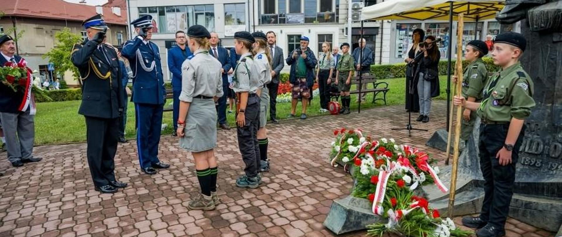 Zdjęcie przedstawia strażaka oraz policjanta, którzy stoją na przed pomnikiem i oddają honory. W tle zgromadzeni ludzie oraz budynki.